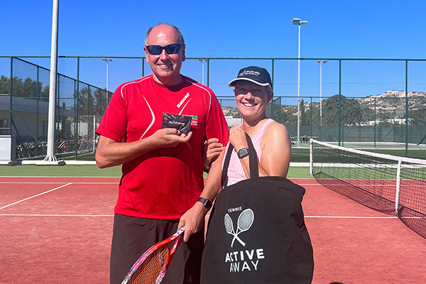 Two tennis players on a red clay court, man in a red shirt holding a racquet, woman with a cap holding a bag labelled 'TENNIS ACTIVE AWAY'.