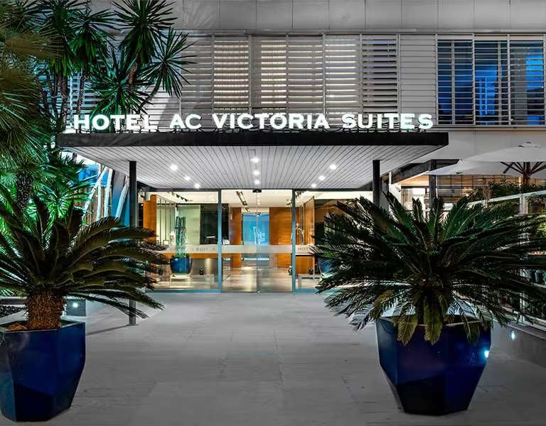 Entrance of Hotel AC Victoria Suites in Barcelona, with glass doors, metal design, and potted palms.