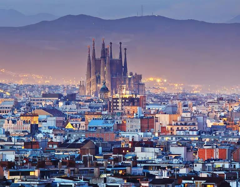 Nighttime image of Barcelona's cityscape featuring the illuminated Sagrada Família basilica, set against a mountainous backdrop.