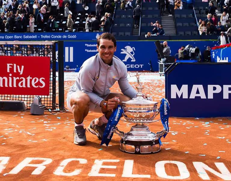 Rafael Nadal kneeling and smiling with the Barcelona Open trophy on a clay court, surrounded by tournament branding and spectators.