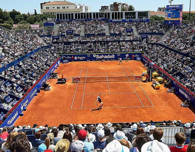 Wide shot of a professional tennis match on a clay court in Barcelona, with a packed stadium of spectators.
