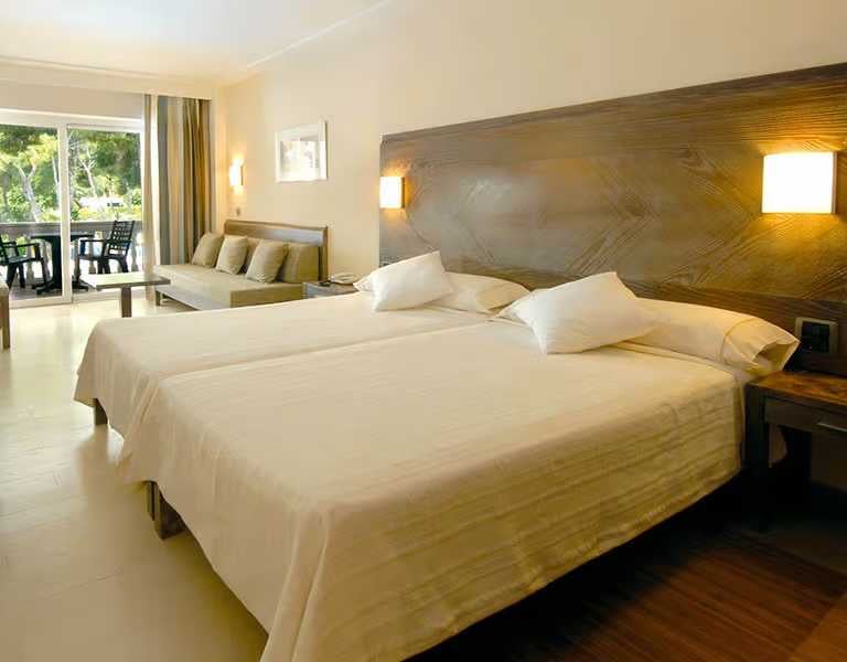 Hotel room with twin beds, seating area, and glass doors opening to a balcony with green views.