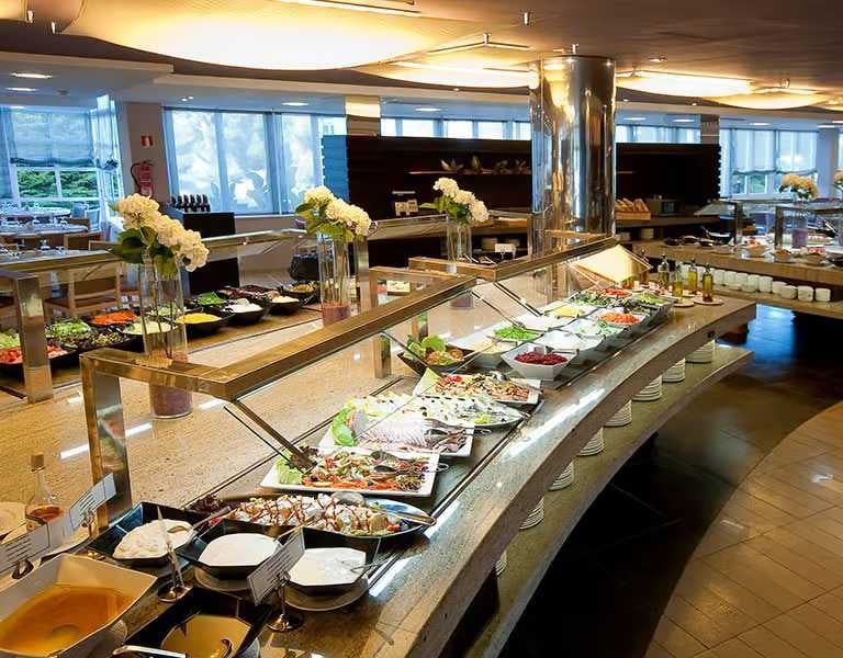 Indoor buffet spread at a beach club with a variety of dishes, salads, platters, and desserts, with decorative flowers and natural light from large windows.