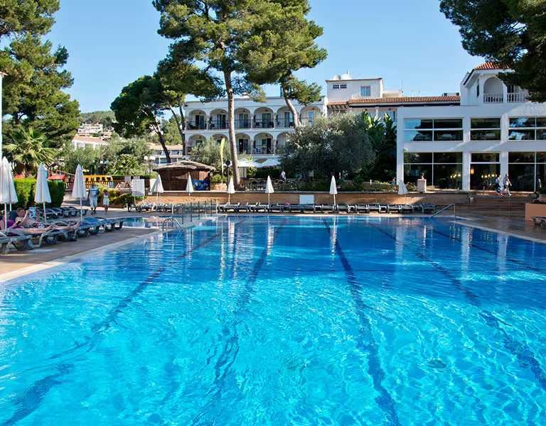 Pool and lounge area at a beach club with sun loungers, parasols, tall trees, and modern white buildings in the background.