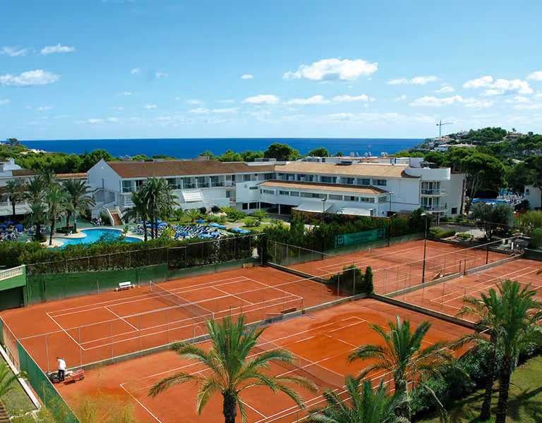 Aerial view of a beach club with clay tennis courts, a pool, white buildings, and sea in the background.