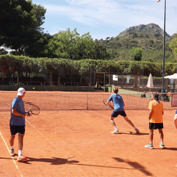 Group of tennis players in a coaching session on a clay court with a coach instructing, set against a backdrop of greenery and mountains.