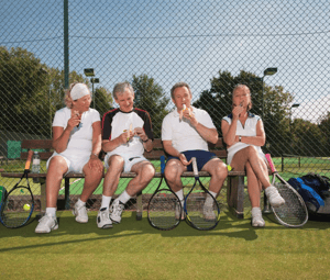 Four adults in tennis gear sitting on a bench by a tennis court, enjoying ice cream cones with rackets and a sports bag nearby.