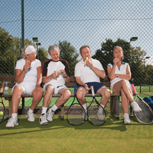 Four adults in tennis gear sitting on a bench by a tennis court, enjoying ice cream cones with rackets and a sports bag nearby.