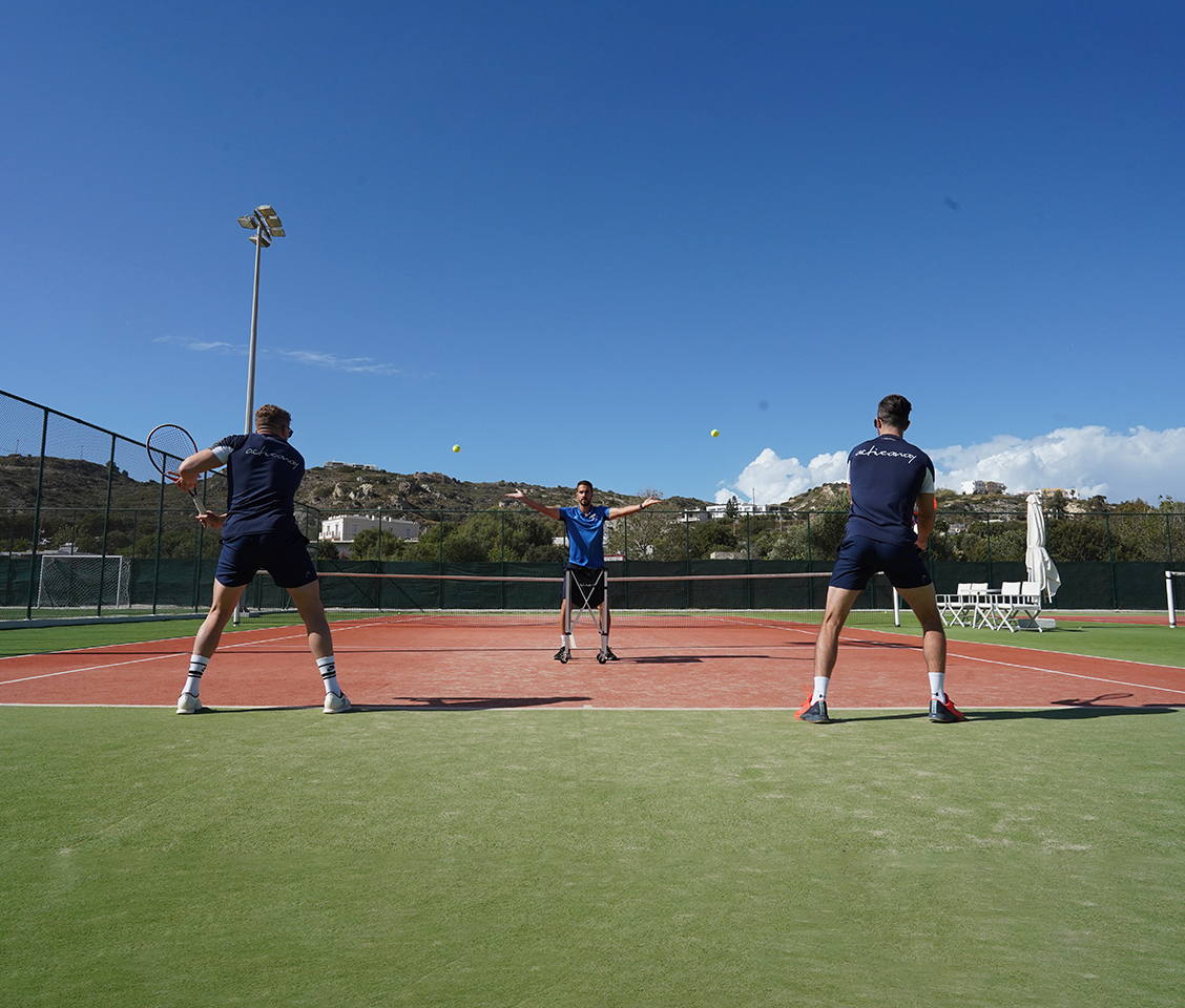 Outdoor tennis coaching session with a coach serving to two players on a clay court under a clear blue sky.