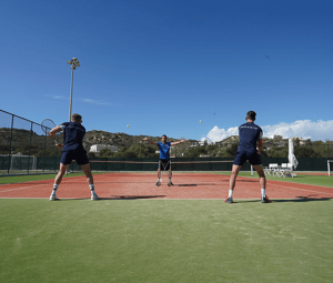 Outdoor tennis coaching session with a coach serving to two players on a clay court under a clear blue sky.