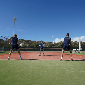 Outdoor tennis coaching session with a coach serving to two players on a clay court under a clear blue sky.