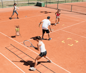 Tennis training session on a clay court with four players, including two women and one man, practising with agility ladders and tennis balls.