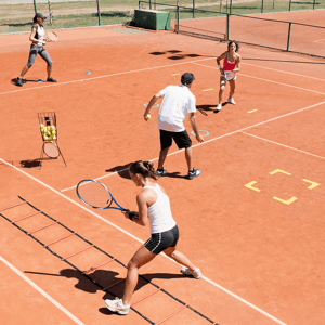 Tennis training session on a clay court with four players, including two women and one man, practising with agility ladders and tennis balls.