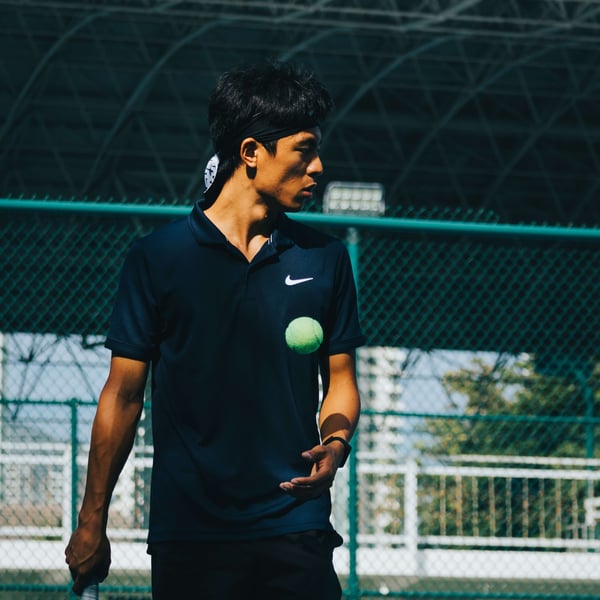 Young male tennis player holding a tennis ball and racket on an outdoor court with a green fence and metal roof behind him.