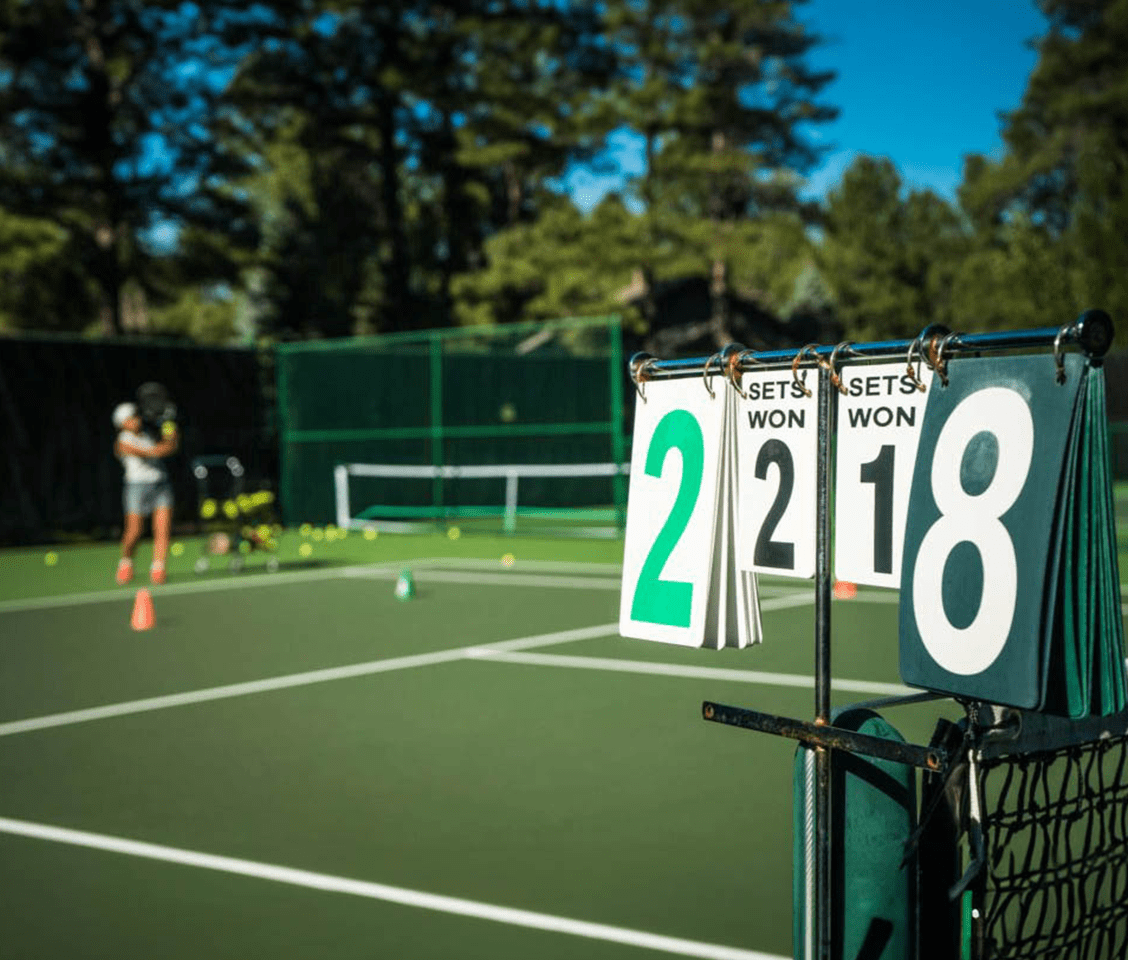 Tennis court with a scorecard showing 2-2 sets, person practising in the background with tennis balls on the court.