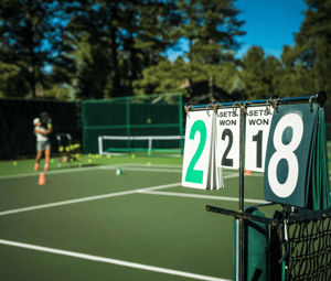 Tennis court with a scorecard showing 2-2 sets, person practising in the background with tennis balls on the court.