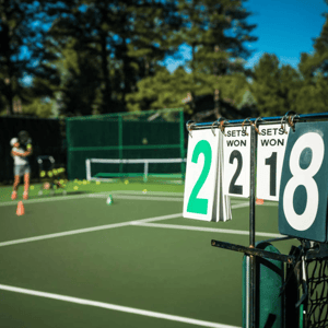 Tennis court with a scorecard showing 2-2 sets, person practising in the background with tennis balls on the court.