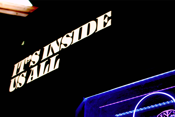 Neon sign reading 'IT'S INSIDE US ALL' with purple and blue lighting on a dark background.