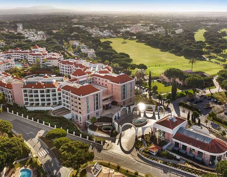 Aerial view of Hilton Vilamoura As Cascatas Golf Resort & Spa, featuring the hotel buildings with red-tiled roofs and an adjacent golf course in lush green surroundings.