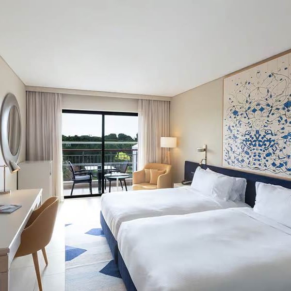 Hilton Vilamoura hotel room with twin beds, abstract wall art, a desk, TV, and a balcony overlooking greenery.