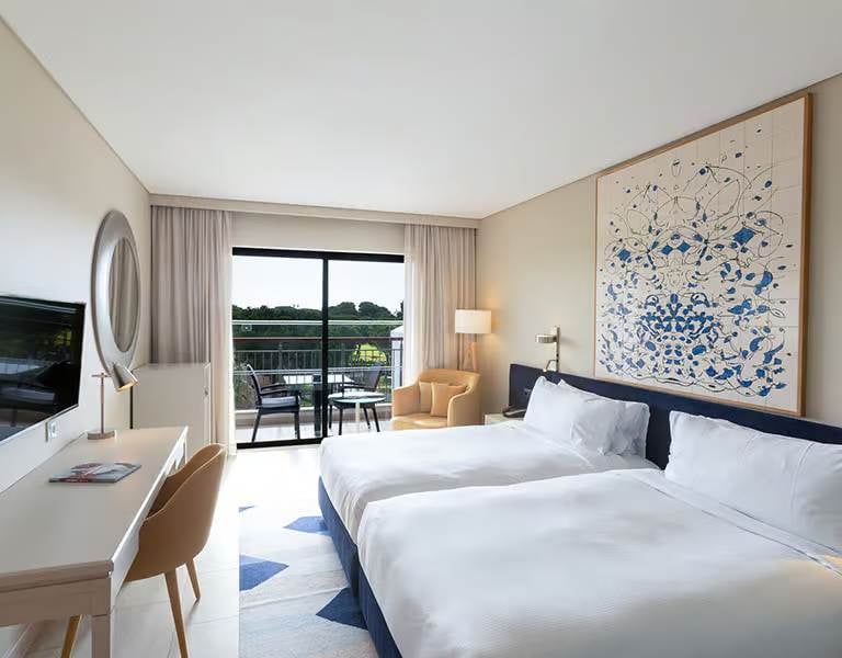 Hilton Vilamoura hotel room with twin beds, abstract wall art, a desk, TV, and a balcony overlooking greenery.