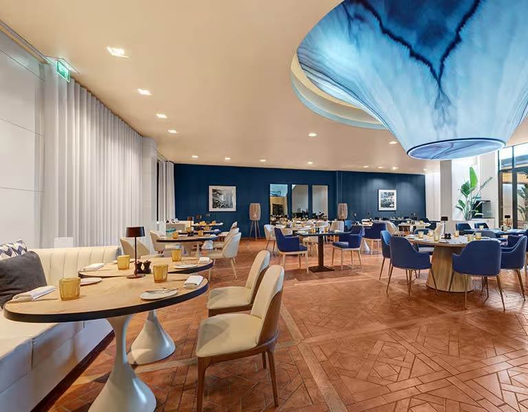 Indoor dining area at Hilton Vilamoura showcasing elegant decor with blue and beige chairs, parquet flooring, and a unique blue wave ceiling fixture.