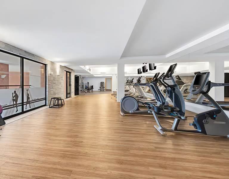 Spacious indoor gym at Hilton Vilamoura with modern exercise equipment including treadmills and elliptical trainers, featuring large windows and wooden flooring.