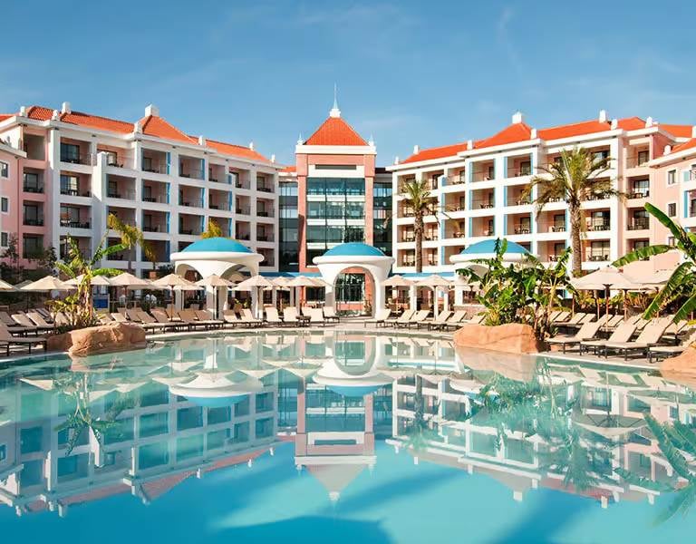 Wide shot of Hilton Vilamoura resort, featuring the hotel exterior with a grand swimming pool and sun loungers in the foreground.