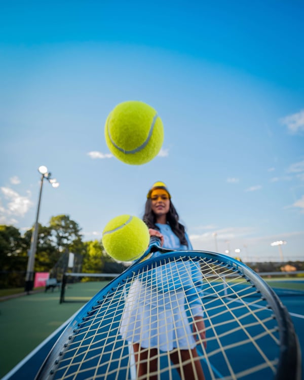 Female tennis player poised with two tennis balls above her racket on an outdoor court under a clear blue sky.