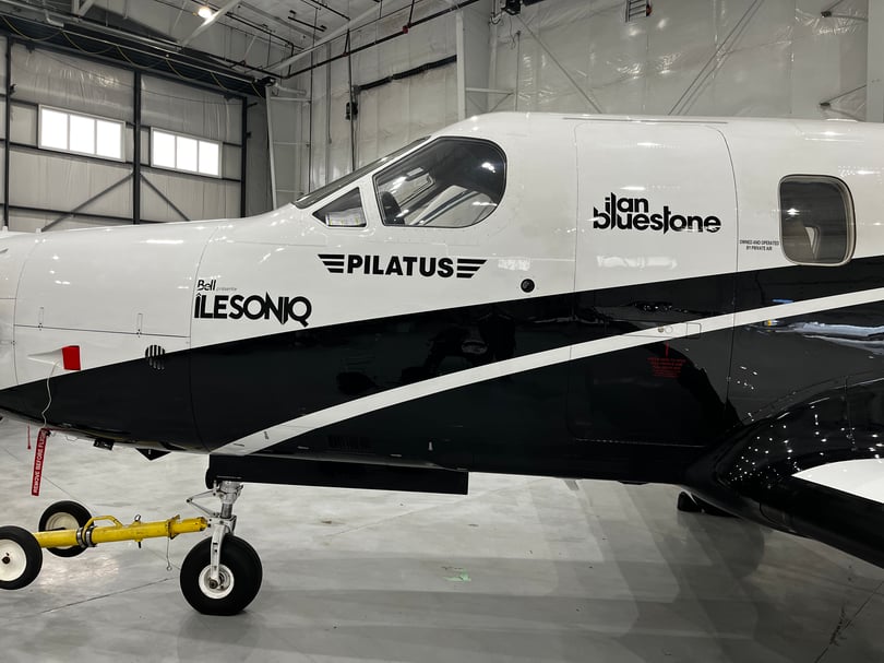 Black sponsor logos and lettering on the white fuselage of a Pilatus aircraft in an indoor hangar