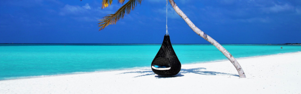 A black hammok chair hanging from a palm tree on a white sandy beach