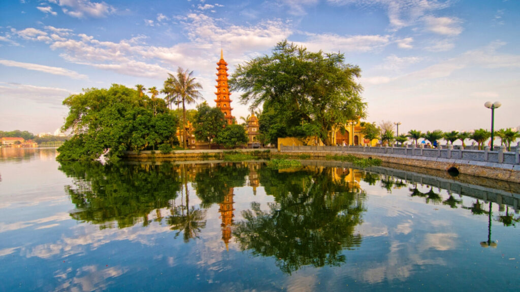 A tall tower of a temple with green trees around and a river in front in Hanoi