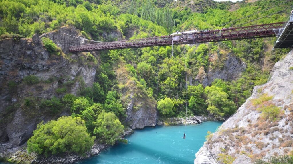 A person bungtjumping from a bridge with a blue river underneath and green nature around