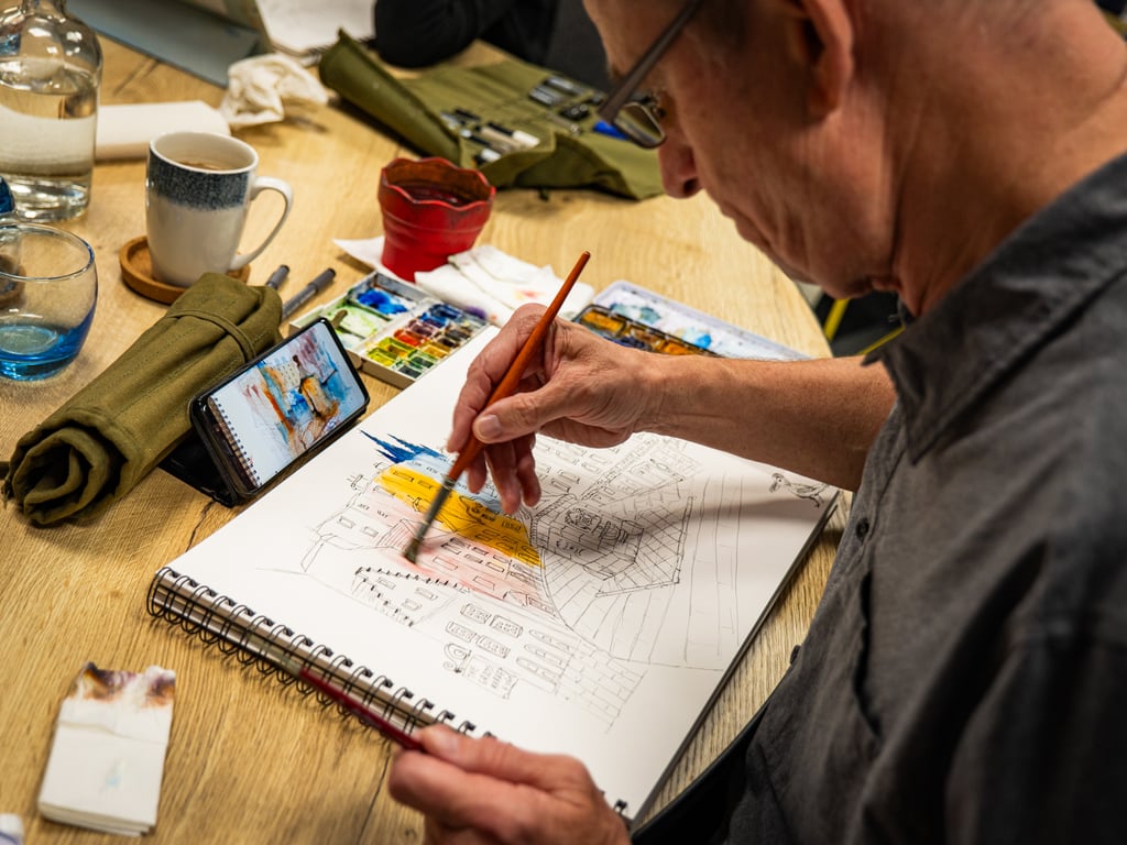 A middle-aged man with salt-and-pepper hair and rectangular glasses is actively sketching at a wooden table during an urban sketching workshop or lesson. He holds a red/brown brush in his hand, adding watercolor washes to a detailed architectural sketch with pen linework. The table is scattered with art materials including a ceramic mug, water glasses, rolled sketching paper, color reference charts on a phone, and a spiral-bound sketchbook. The warm, collaborative atmosphere suggests an instructional or mentoring setting where sketching techniques are being demonstrated and practiced.
