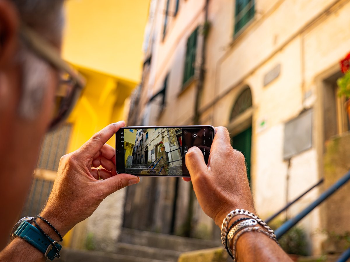 Hands holding a phone up to photograph a narrow sunlit street, the framed scene visible on the screen.