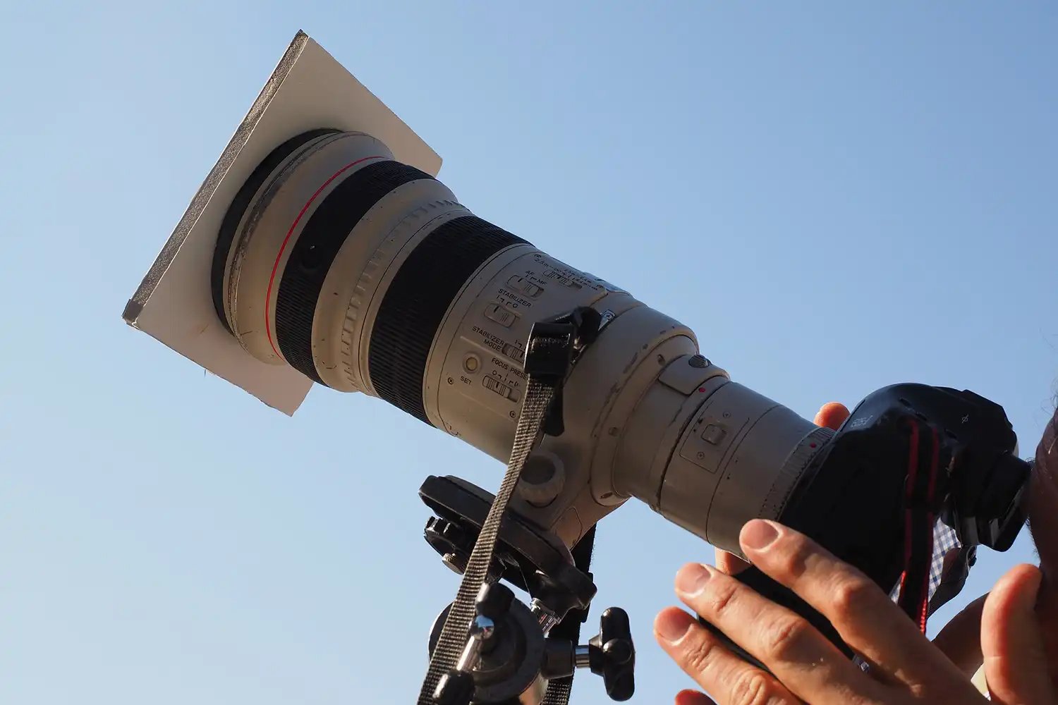 a Canon DSLR camera with telephoto lens pointing up to the sky with a cardboard cover over the lens for shooting a solar eclipse