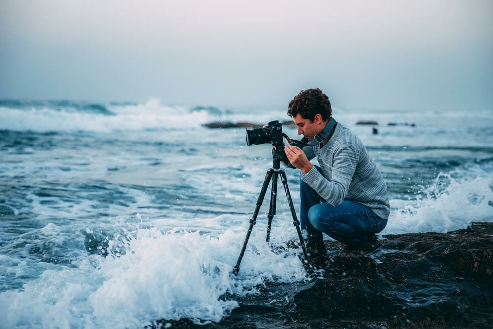 Young handsome photographer with curly hair, a gray sweater and jeans with a tripod and a camera on the beach, drenched in waves. Photography in Israel. Copy space. Shtorm on Mediterranean Sea