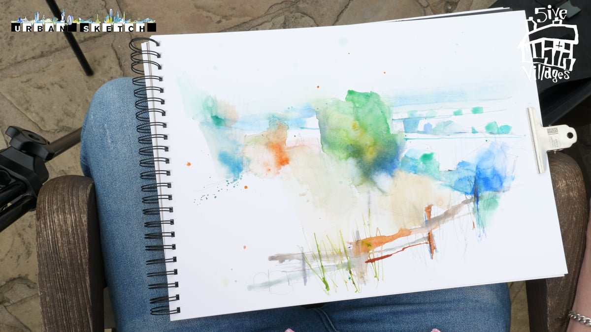 A watercolor sketch in progress displayed on a spiral-bound sketchbook resting on the artist's lap. The loose, expressionistic landscape features vibrant washes of green, blue, orange, and yellow with architectural elements and foliage suggested through flowing watercolor blooms. The outdoor setting shows the sketch from an overhead perspective with natural daylight, capturing the spontaneous, fluid nature of urban watercolor painting in the field.