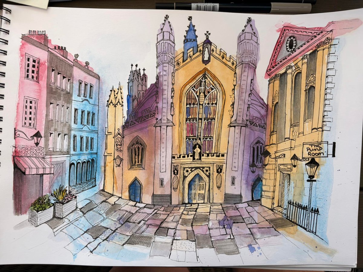 A detailed watercolor and ink urban sketch in a spiral-bound sketchbook depicting a charming European street scene centered on a grand Gothic church. The composition features the ornate church with golden-yellow and purple spires at the center, flanked by colorful residential buildings in soft pink, blue, and cream tones with detailed architectural elements like balconies and windows. The foreground shows an intricate tiled street floor rendered in perspective with purple, pink, and gray hues. The sketch combines architectural precision with expressive, gestural linework and vibrant watercolor washes, creating a lively, atmospheric mood that captures the character of a historic European city street.