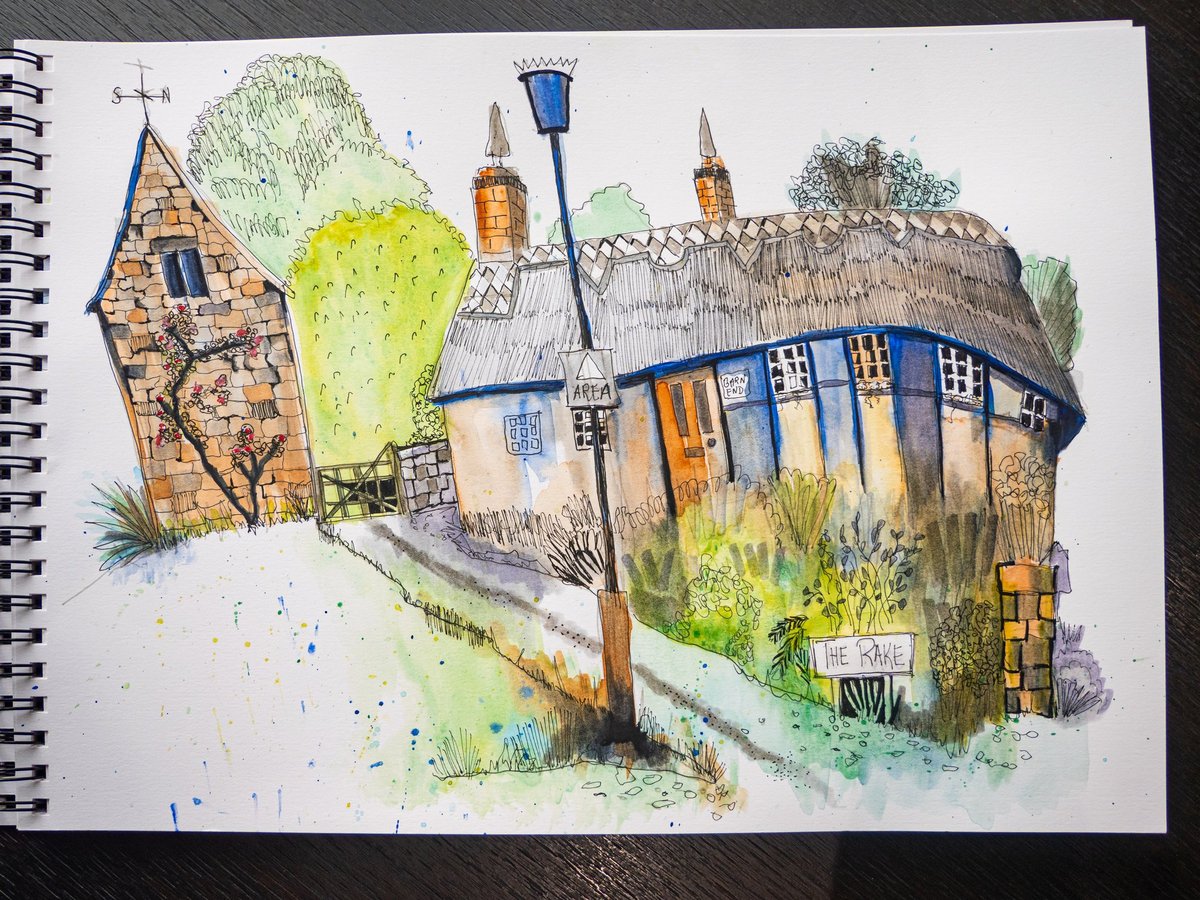 A beautifully detailed urban sketch in pen and watercolor showing a charming English country cottage scene. The composition features two distinctive thatched-roof buildings with stone/brick walls in warm orange and tan tones, complete with blue doors and windows. Lush green foliage surrounds the structures, with blue lamp post and directional signage visible. The sketch demonstrates skilled linework with fine ink details combined with vibrant watercolor washes in greens, yellows, and blues, with splashes of color suggesting a spontaneous, artistic approach.