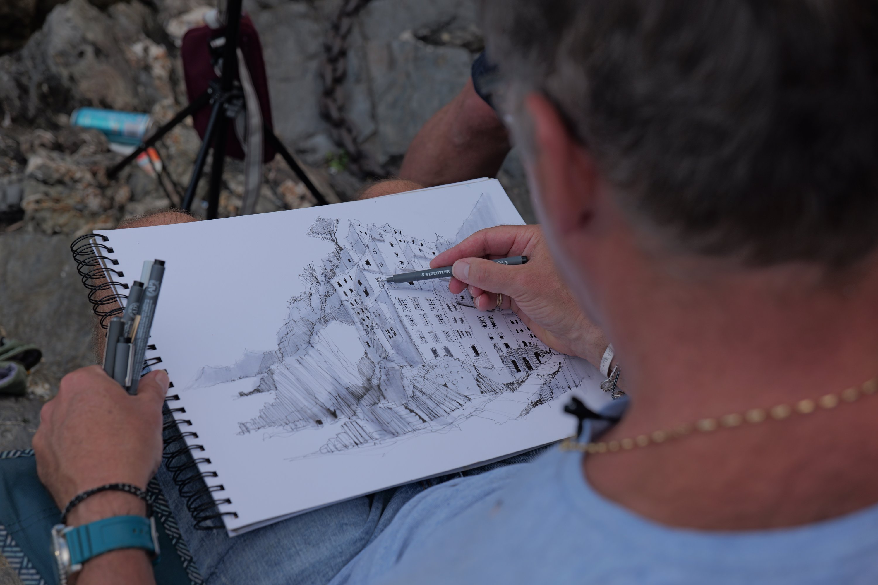Artist sketching a hilltop village in pen on a spiral-bound sketchbook, viewed over their shoulder on location