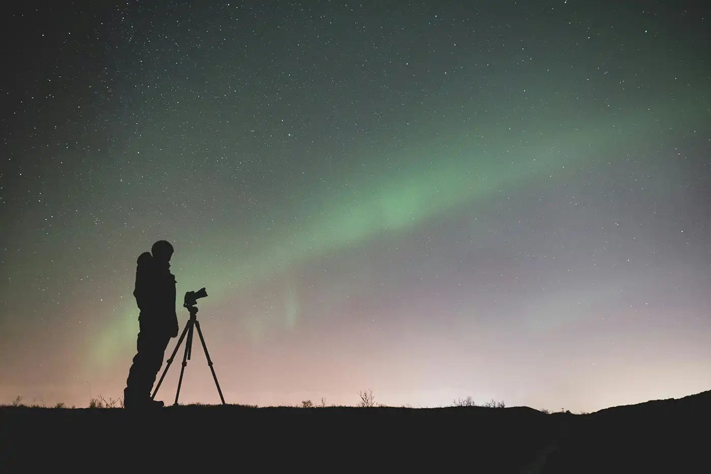 a man standing in silhouette in front of a camera on a tripod taking photos of the northern lights