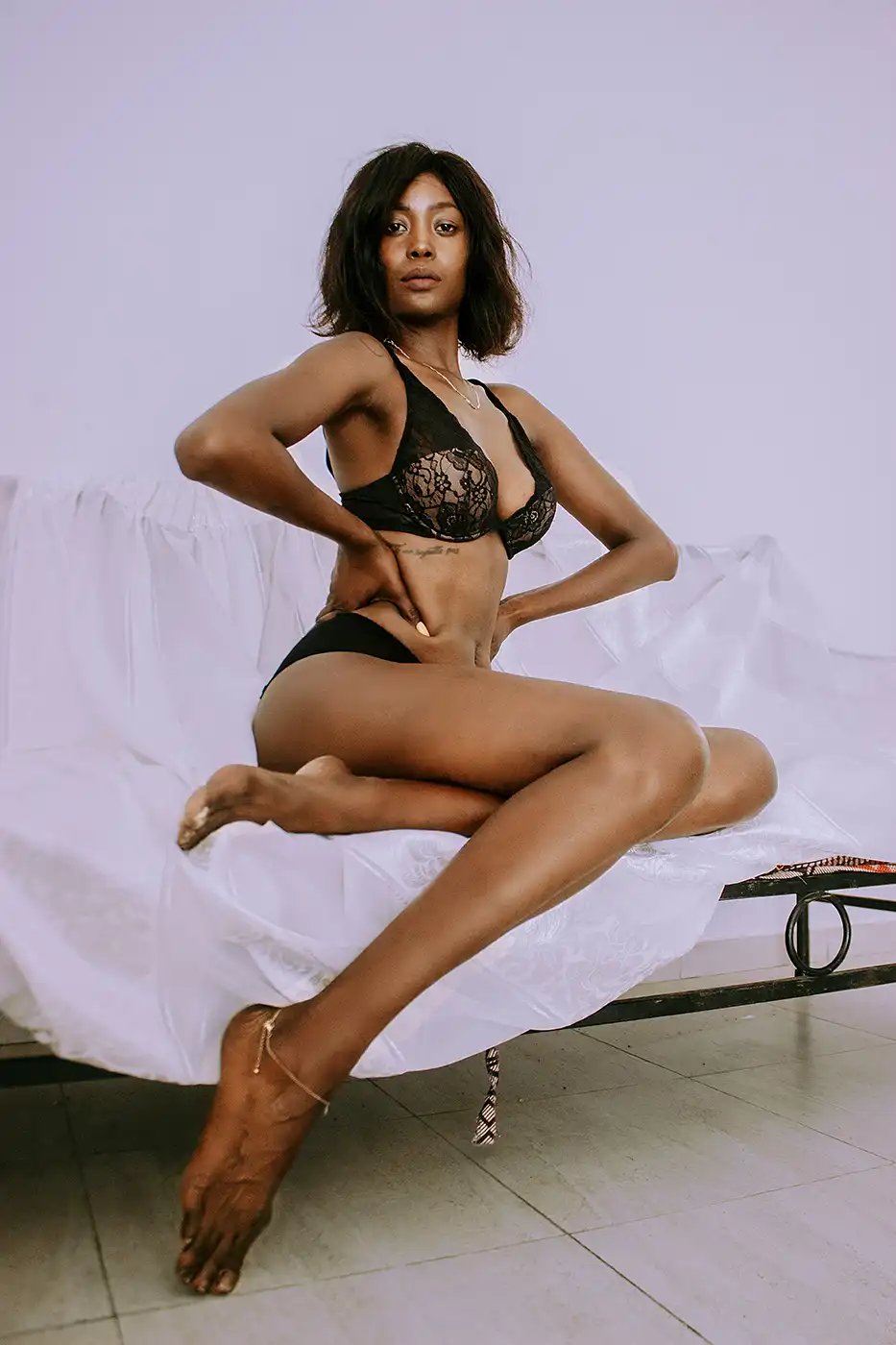 a black woman wearing black underwear hands on hips posing on the end of a white covered chair