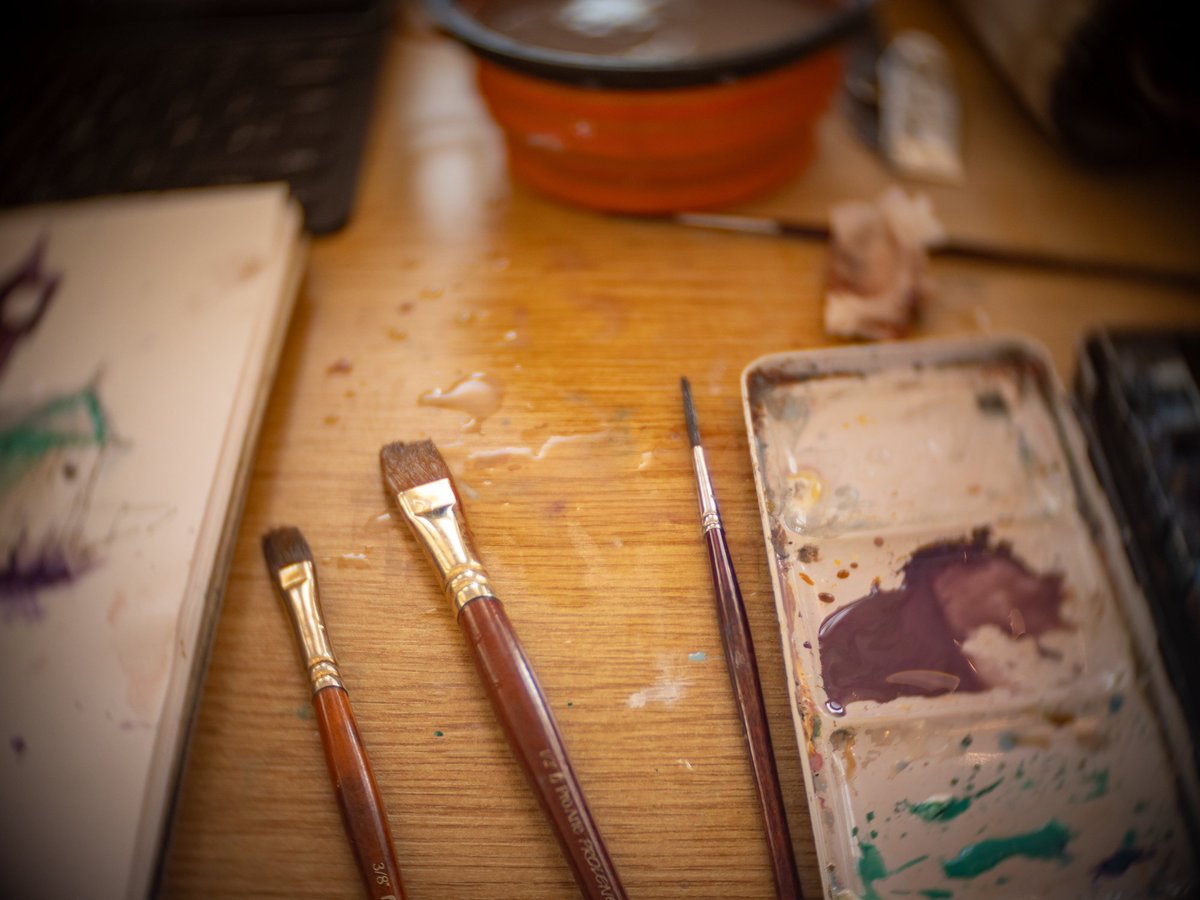 An artist's working setup captured from above, showing three paintbrushes with wooden handles and worn brass ferrules arranged on a honey-toned wooden surface. A ceramic palette with dried paint residue in earth tones (browns, greens) sits to the right, while sketching materials and an orange ceramic bowl are visible blurred in the background. The warm, natural lighting and paint-splattered surfaces evoke an active creative workspace.