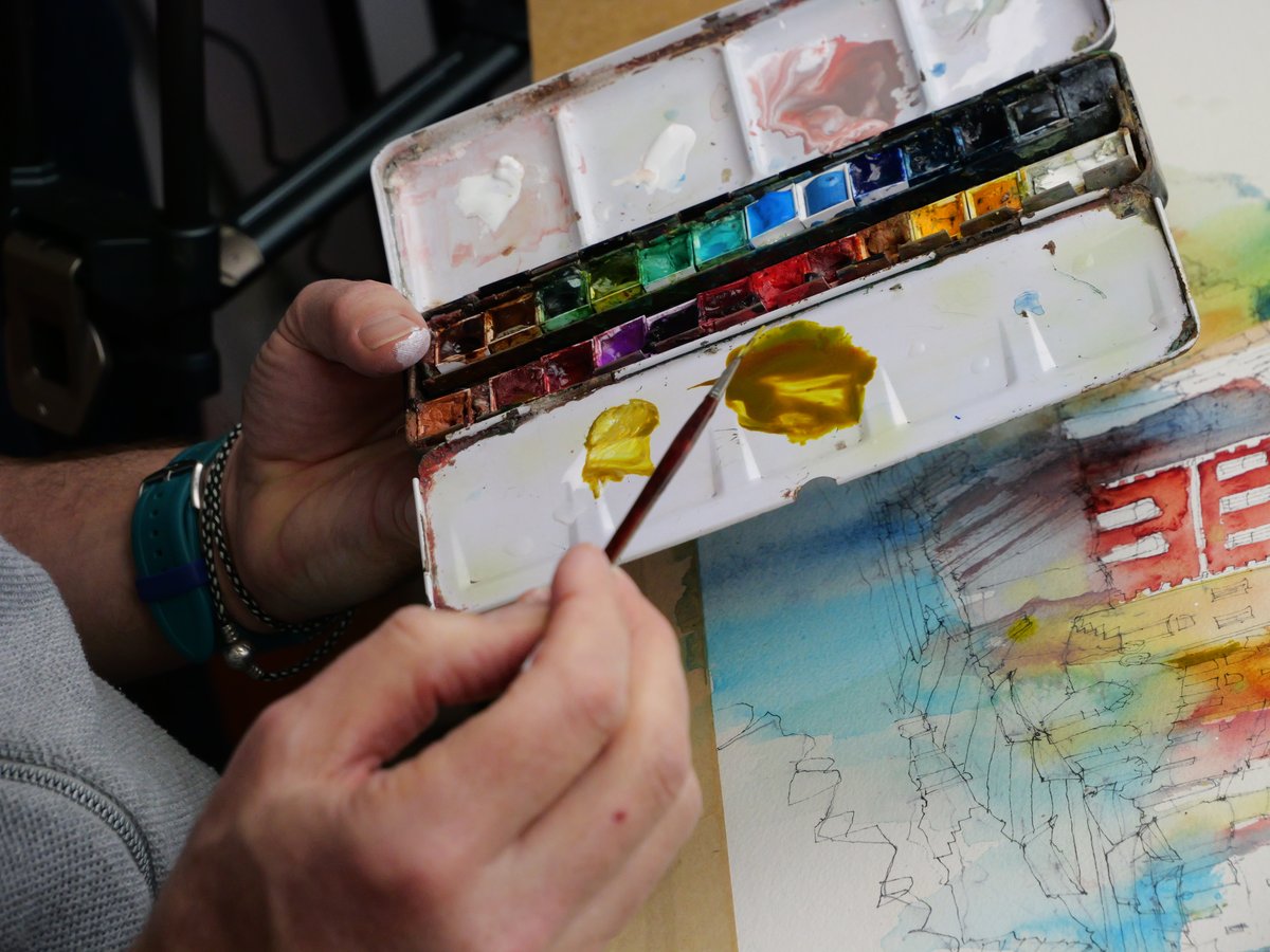 A close-up shot of hands holding an open watercolor palette with vibrant pigment blocks in blue, yellow, orange, red, purple, and green. The artist is actively painting with a wet brush, with bright yellow watercolor applied to the palette. Below the palette is a partially completed urban sketch featuring architectural linework, red building details, and watercolor washes in blue, yellow, and warm tones. The artist wears bracelets and appears to be working outdoors or in a fieldwork setting.