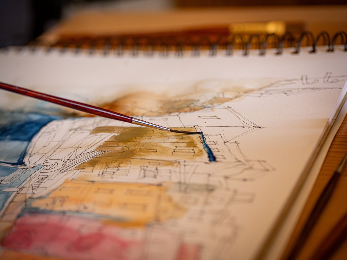 A close-up view of an open sketchbook with an in-progress urban sketch featuring architectural drawings with colored washes in ochre, blue, and pink. A dark red paintbrush hovers over the work, actively applying watercolor. A spiral-bound notebook is visible in the soft-focus background, and the entire scene is lit warmly, creating a natural studio atmosphere on a wooden work surface.