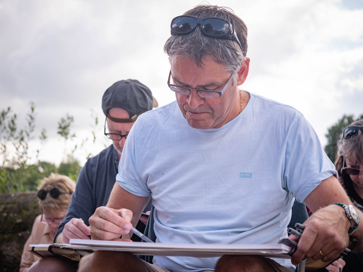 A middle-aged man in a light blue t-shirt sketches outdoors in an open natural setting, surrounded by attentive students in a group learning environment. He wears glasses pushed up on his forehead and appears focused on his sketchpad while participants gather around him. The scene captures an instructor-led sketching workshop with a cloudy sky and green vegetation visible in the background, suggesting an outdoor field teaching moment.