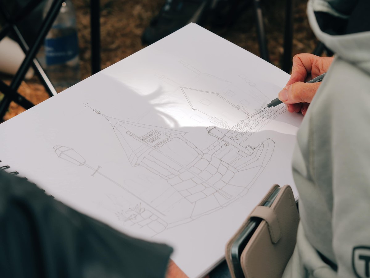 A hands-on sketching moment outdoors showing a person actively drawing detailed architectural line sketches on white paper. The sketch depicts building interiors and structural elements with precise pencil work. The scene captures the intimate creative process in natural daylight with blurred background elements including folding chairs and outdoor furniture, suggesting an outdoor art class or plein air sketching session.