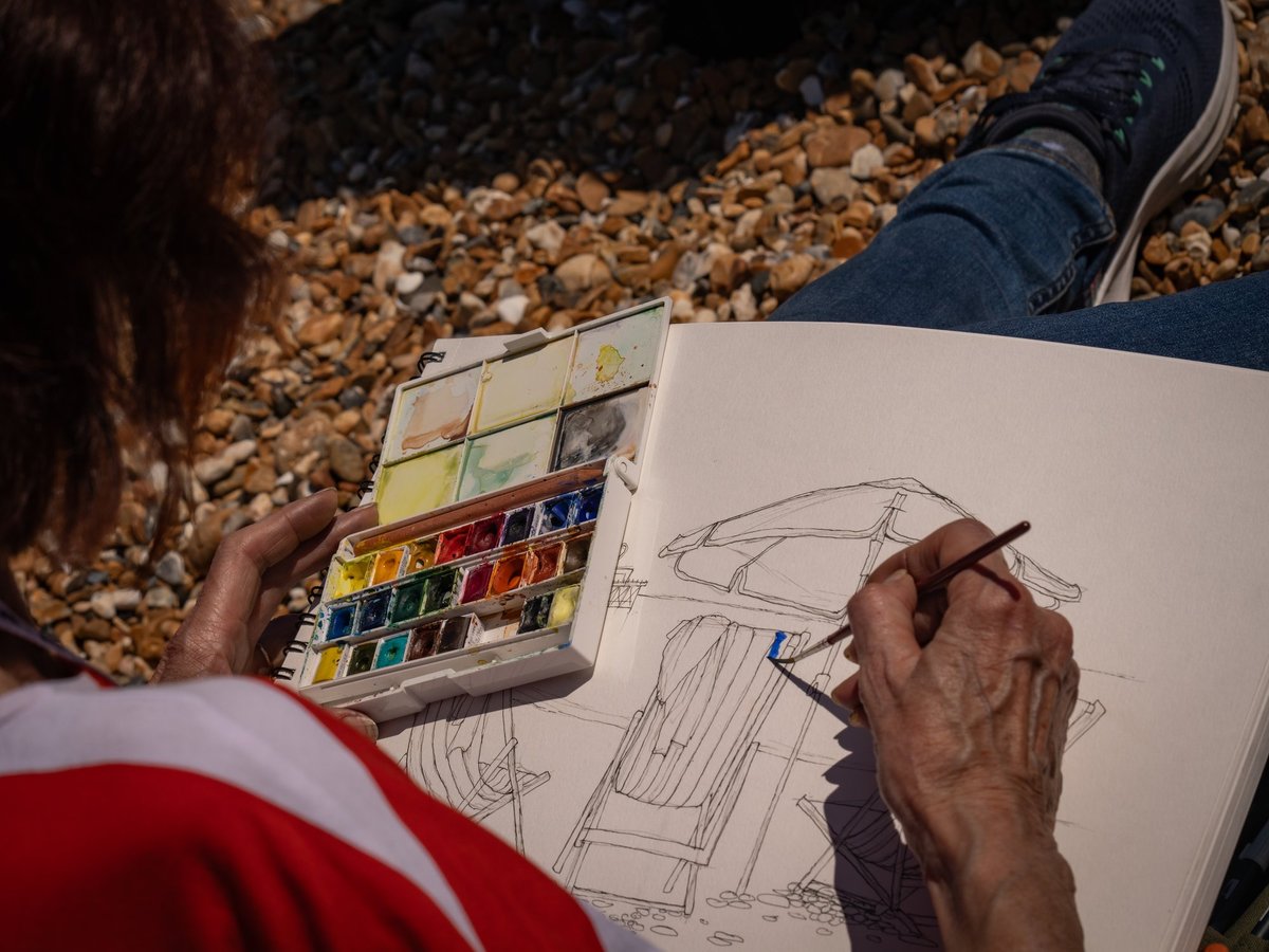 A sketcher in progress sits outdoors on a pebbly beach, holding an open portable watercolor palette and actively painting architectural line drawings on white paper. The artist wears a red and white jacket and blue jeans, with the beach pebbles (golden, tan, and white stones) clearly visible beneath them. The sketch appears to show building or street scenes rendered in ink with blue watercolor accents, demonstrating on-location artistic practice with natural light and minimal materials.
