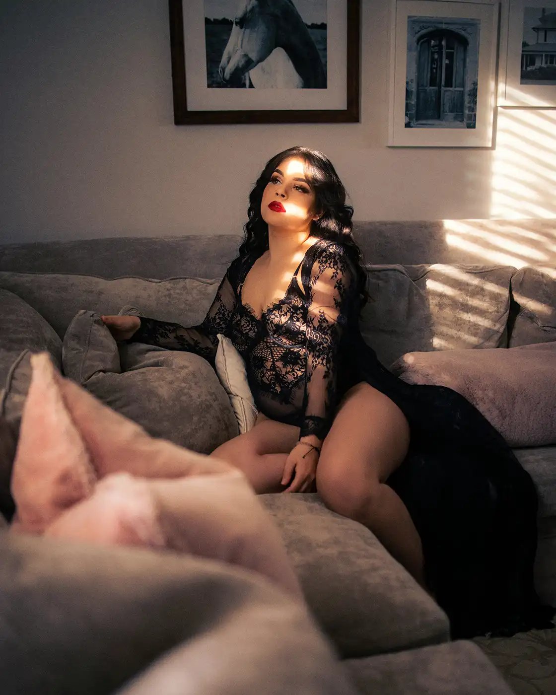 A woman in a black lace dress looking out a window sat on a sofa